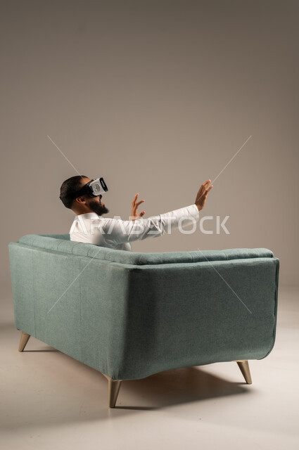 قضاء أوقات ممتعة ومسلية بالألعاب الافتراضية، الاستمتاع بمشاهدة أفلام الواقع الافتراضي، بورتريه من الخلف لرجل عربي خليجي سعودي يرتدي الثوب التقليدي يجلس على كرسي مريح ويرتدي نظارة الواقع الافتراضي ثلاثية الابعاد، خلفية رمادية