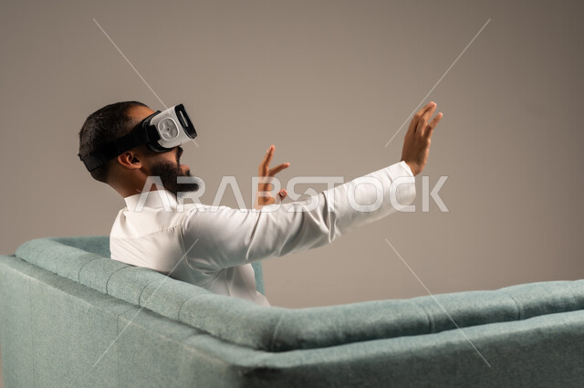 قضاء أوقات ممتعة ومسلية بالألعاب الافتراضية، الاستمتاع بمشاهدة أفلام الواقع الافتراضي، بورتريه من الخلف لرجل عربي خليجي سعودي يرتدي الثوب التقليدي يجلس على كرسي مريح ويرتدي نظارة الواقع الافتراضي ثلاثية الابعاد، خلفية رمادية