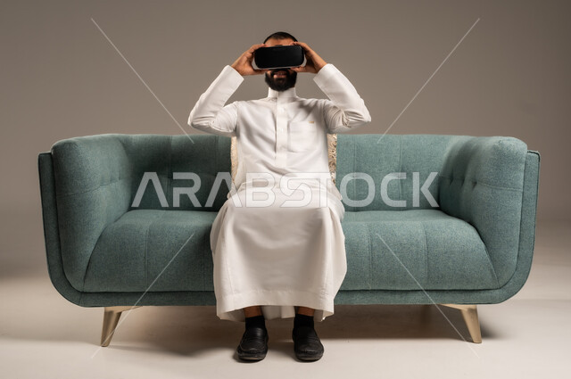 الاستمتاع بمشاهدة أفلام الواقع الافتراضي، قضاء أوقات ممتعة ومسلية بالألعاب الافتراضية،  بورتريه لرجل عربي خليجي سعودي يرتدي الثوب التقليدي يجلس على كرسي مريح ويرتدي نظارة الواقع الافتراضي ثلاثية الابعاد، خلفية رمادية