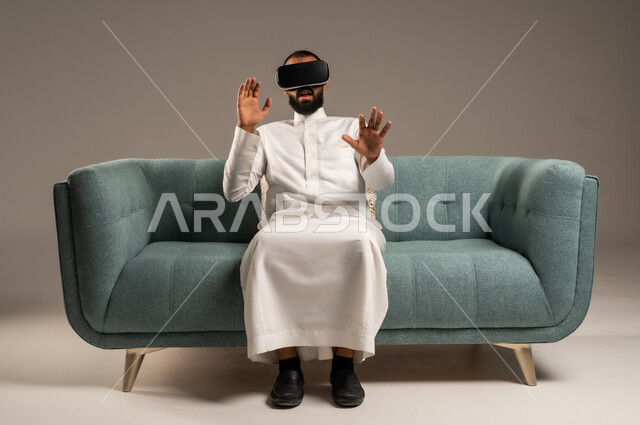 الاستمتاع بمشاهدة أفلام الواقع الافتراضي، قضاء أوقات ممتعة ومسلية بالألعاب الافتراضية،  بورتريه لرجل عربي خليجي سعودي يرتدي الثوب التقليدي يجلس على كرسي مريح ويرتدي نظارة الواقع الافتراضي ثلاثية الابعاد، خلفية رمادية