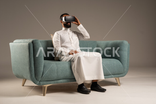 الاستمتاع بمشاهدة أفلام الواقع الافتراضي، قضاء أوقات ممتعة ومسلية بالألعاب الافتراضية،  بورتريه لرجل عربي خليجي سعودي يرتدي الثوب التقليدي يجلس على كرسي مريح ويرتدي نظارة الواقع الافتراضي ثلاثية الابعاد، خلفية رمادية