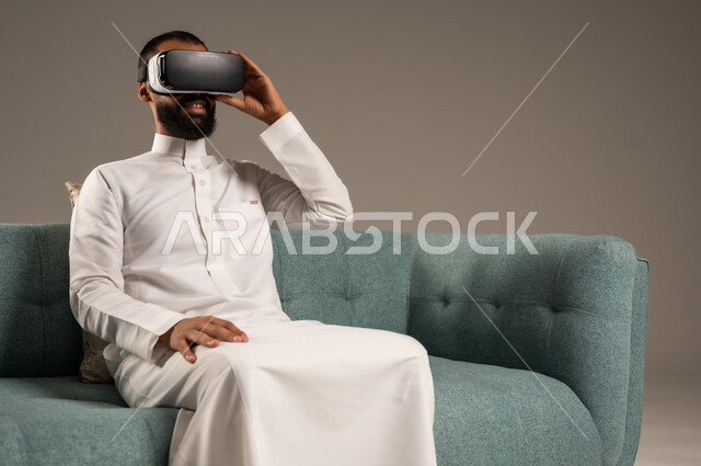 الاستمتاع بمشاهدة أفلام الواقع الافتراضي، قضاء أوقات ممتعة ومسلية بالألعاب الافتراضية،  بورتريه لرجل عربي خليجي سعودي يرتدي الثوب التقليدي يجلس على كرسي مريح ويرتدي نظارة الواقع الافتراضي ثلاثية الابعاد، خلفية رمادية