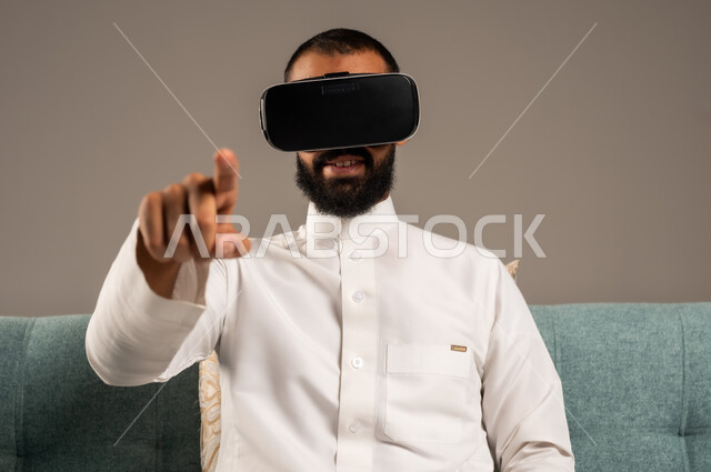 الاندماج ومحاكاة العالم الخيالي، الاستمتاع بمشاهدة أفلام الواقع الافتراضي، قضاء أوقات ممتعة ومسلية بالألعاب الافتراضية،  بورتريه لرجل عربي خليجي سعودي يرتدي الثوب التقليدي يجلس على كرسي مريح ويرتدي نظارة الواقع الافتراضي ثلاثية الابعاد، خلفية رمادية