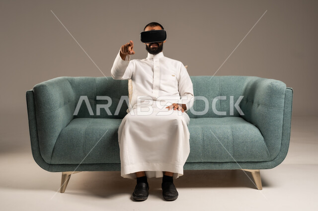 Integration and simulation of the imaginary world, enjoying watching virtual reality movies, spending fun and entertaining times with virtual games, portrait of a Saudi Arabian Gulf man wearing traditional dress sitting on a comfortable chair and wearing 3D virtual reality glasses, gray background