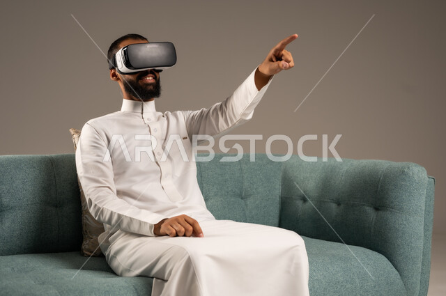 الاندماج ومحاكاة العالم الخيالي، الاستمتاع بمشاهدة أفلام الواقع الافتراضي، قضاء أوقات ممتعة ومسلية بالألعاب الافتراضية،  بورتريه لرجل عربي خليجي سعودي يرتدي الثوب التقليدي يجلس على كرسي مريح ويرتدي نظارة الواقع الافتراضي ثلاثية الابعاد، خلفية رمادية