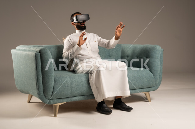 الاندماج ومحاكاة العالم الخيالي، الاستمتاع بمشاهدة أفلام الواقع الافتراضي، قضاء أوقات ممتعة ومسلية بالألعاب الافتراضية،  بورتريه لرجل عربي خليجي سعودي يرتدي الثوب التقليدي يجلس على كرسي مريح ويرتدي نظارة الواقع الافتراضي ثلاثية الابعاد، خلفية رمادية