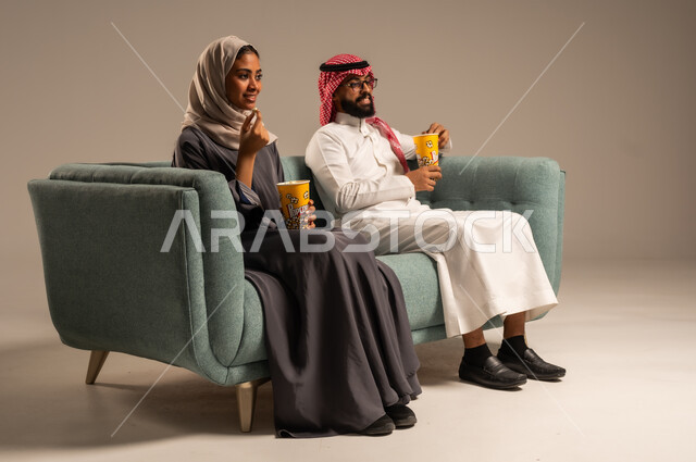 Enjoying family fun activities on the weekend, having a good time at home, portrait of a Saudi Arabian Gulf couple sitting on a comfortable chair in the living room eating popcorn, loving atmosphere, watching movies and series, gray background