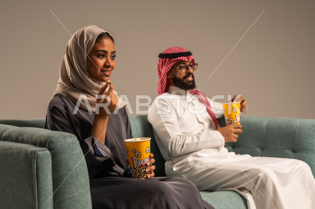  الاستمتاع بالأنشطة العائلية الترفيهيه في عطلة نهاية الاسبوع, قضاء وقت ممتع في المنزل, بورتريه لزوجان عربيان خليجيان سعوديان يجلسان على كرسي مريح في غرفة المعيشة يقومان بتناول الفوشار، أجواء مليئة بالحب, مشاهدة الأفلام والمسلسلات، خلفية رمادية