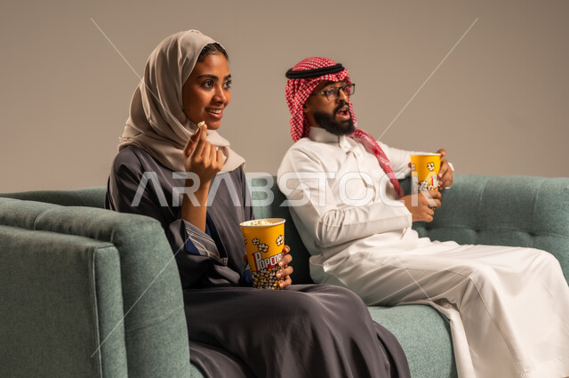  الاستمتاع بالأنشطة العائلية الترفيهيه في عطلة نهاية الاسبوع, قضاء وقت ممتع في المنزل, بورتريه لزوجان عربيان خليجيان سعوديان يجلسان على كرسي مريح في غرفة المعيشة يقومان بتناول الفوشار، أجواء مليئة بالحب, مشاهدة الأفلام والمسلسلات، خلفية رمادية