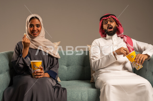  الاستمتاع بالأنشطة العائلية الترفيهيه في عطلة نهاية الاسبوع, قضاء وقت ممتع في المنزل, بورتريه لزوجان عربيان خليجيان سعوديان يجلسان على كرسي مريح في غرفة المعيشة يقومان بتناول الفوشار، أجواء مليئة بالحب, مشاهدة الأفلام والمسلسلات، خلفية رمادية