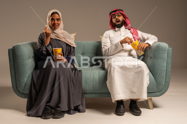  الاستمتاع بالأنشطة العائلية الترفيهيه في عطلة نهاية الاسبوع, قضاء وقت ممتع في المنزل, بورتريه لزوجان عربيان خليجيان سعوديان يجلسان على كرسي مريح في غرفة المعيشة يقومان بتناول الفوشار، أجواء مليئة بالحب, مشاهدة الأفلام والمسلسلات، خلفية رمادية