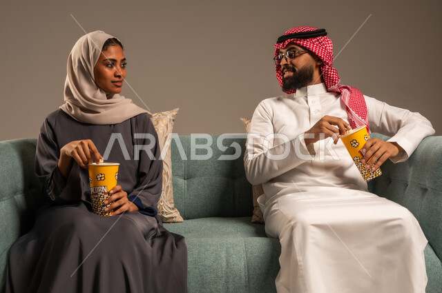  أجواء مليئة بالحب, الاستمتاع بالأنشطة العائلية الترفيهيه في عطلة نهاية الاسبوع, قضاء وقت ممتع في المنزل, بورتريه لزوجان عربيان خليجيان سعوديان يجلسان على كرسي مريح في غرفة المعيشة يقومان بتناول الفوشار، مشاهدة الأفلام والمسلسلات، خلفية رمادية