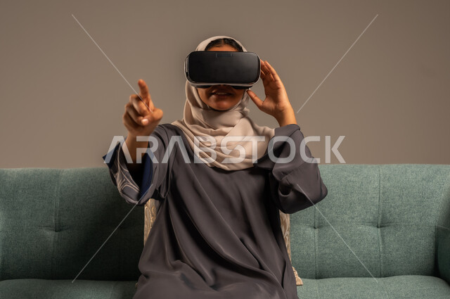 وسائل ترفيهية في وقت الفراغ, الاستمتاع بمشاهدة أفلام الواقع الافتراضي، بورتريه لإمرأة عربية خليجية سعودية تجلس على كرسي مريح وترتدي نظارة الواقع الافتراضي ثلاثية الأبعاد، قضاء أوقات ممتعة ومسلية بالألعاب الافتراضية، خلفية رمادية