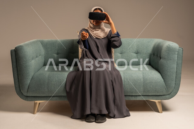وسائل ترفيهية في وقت الفراغ, الاستمتاع بمشاهدة أفلام الواقع الافتراضي، بورتريه لإمرأة عربية خليجية سعودية تجلس على كرسي مريح وترتدي نظارة الواقع الافتراضي ثلاثية الأبعاد، قضاء أوقات ممتعة ومسلية بالألعاب الافتراضية، خلفية رمادية