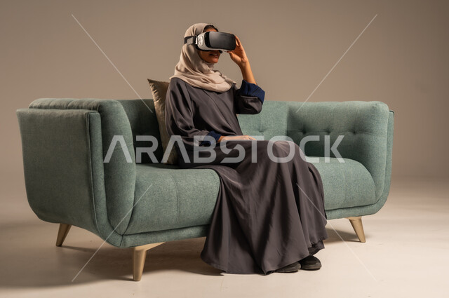وسائل ترفيهية في وقت الفراغ, الاستمتاع بمشاهدة أفلام الواقع الافتراضي، بورتريه لإمرأة عربية خليجية سعودية تجلس على كرسي مريح وترتدي نظارة الواقع الافتراضي ثلاثية الأبعاد، قضاء أوقات ممتعة ومسلية بالألعاب الافتراضية، خلفية رمادية