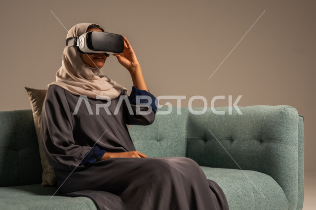 وسائل ترفيهية في وقت الفراغ, الاستمتاع بمشاهدة أفلام الواقع الافتراضي، بورتريه لإمرأة عربية خليجية سعودية تجلس على كرسي مريح وترتدي نظارة الواقع الافتراضي ثلاثية الأبعاد، قضاء أوقات ممتعة ومسلية بالألعاب الافتراضية، خلفية رمادية
