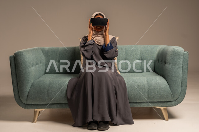 الاستمتاع بمشاهدة أفلام الواقع الافتراضي، وسائل ترفيهية في وقت الفراغ,  بورتريه لإمرأة عربية خليجية سعودية تجلس على كرسي مريح وترتدي نظارة الواقع الافتراضي ثلاثية الأبعاد، قضاء أوقات ممتعة ومسلية بالألعاب الافتراضية، خلفية رمادية