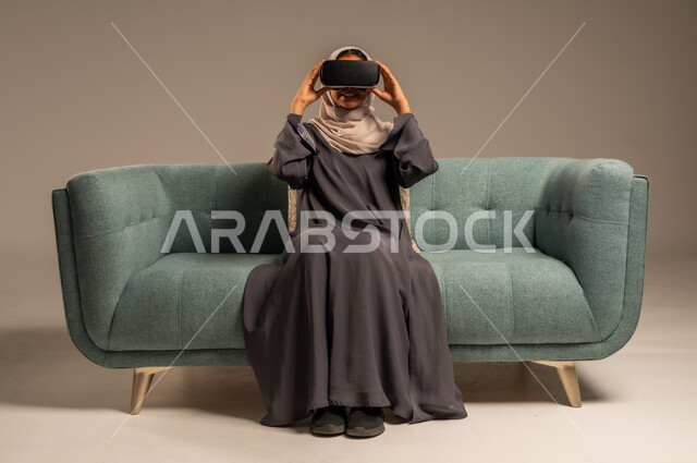 الاستمتاع بمشاهدة أفلام الواقع الافتراضي، وسائل ترفيهية في وقت الفراغ,  بورتريه لإمرأة عربية خليجية سعودية تجلس على كرسي مريح وترتدي نظارة الواقع الافتراضي ثلاثية الأبعاد، قضاء أوقات ممتعة ومسلية بالألعاب الافتراضية، خلفية رمادية