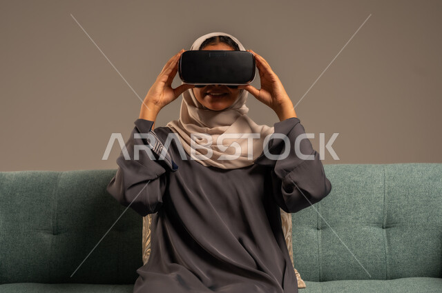الاستمتاع بمشاهدة أفلام الواقع الافتراضي، وسائل ترفيهية في وقت الفراغ,  بورتريه لإمرأة عربية خليجية سعودية تجلس على كرسي مريح وترتدي نظارة الواقع الافتراضي ثلاثية الأبعاد، قضاء أوقات ممتعة ومسلية بالألعاب الافتراضية، خلفية رمادية