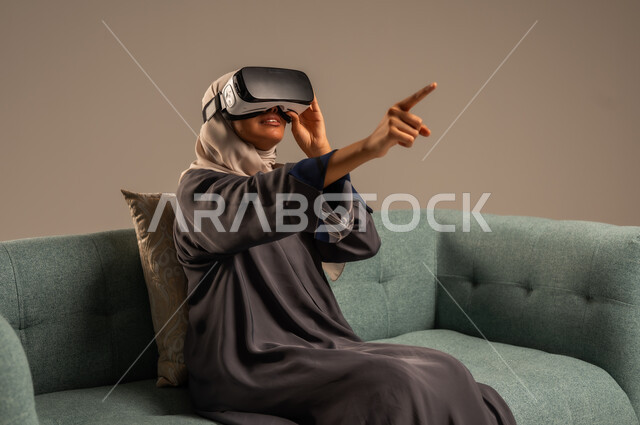 الاستمتاع بمشاهدة أفلام الواقع الافتراضي، وسائل ترفيهية في وقت الفراغ,  بورتريه لإمرأة عربية خليجية سعودية تجلس على كرسي مريح وترتدي نظارة الواقع الافتراضي ثلاثية الأبعاد، قضاء أوقات ممتعة ومسلية بالألعاب الافتراضية، خلفية رمادية