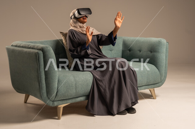 مشاهدة أفلام الواقع الافتراضي، وسائل ترفيهية في وقت الفراغ,  بورتريه لإمرأة عربية خليجية سعودية تجلس على كرسي مريح وترتدي نظارة الواقع الافتراضي ثلاثية الأبعاد، قضاء أوقات ممتعة ومسلية بالألعاب الافتراضية، خلفية رمادية