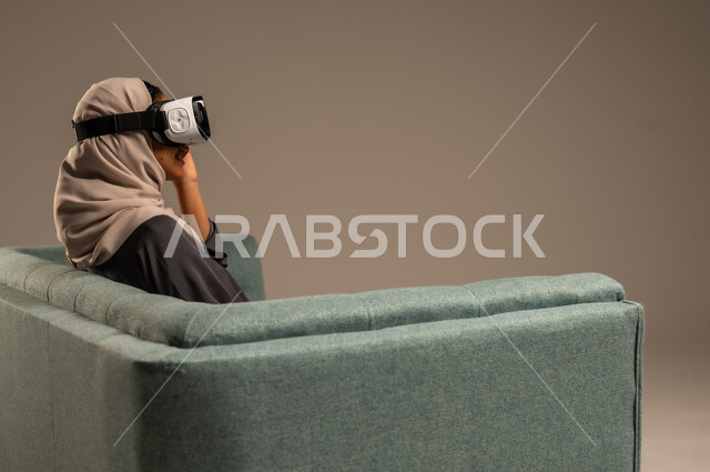 Watching virtual reality movies, leisure time entertainment, portrait of a Saudi Arabian Gulf woman sitting on a comfortable chair wearing 3D virtual reality glasses, having fun and entertaining times with virtual games, gray background