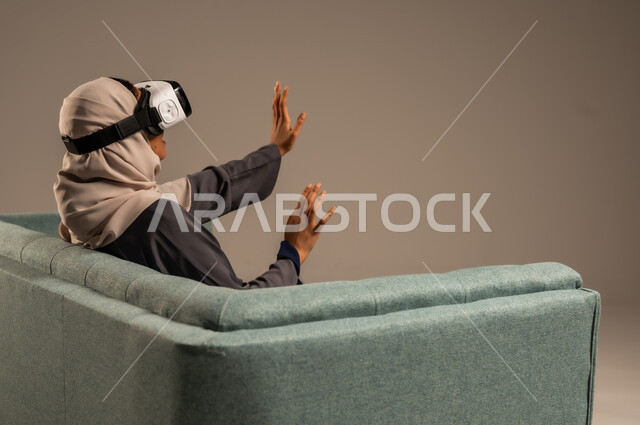 قضاء أوقات ممتعة ومسلية بالألعاب الافتراضية، مشاهدة أفلام الواقع الافتراضي، وسائل ترفيهية في وقت الفراغ,  بورتريه لإمرأة عربية خليجية سعودية تجلس على كرسي مريح وترتدي نظارة ثلاثية الأبعاد، خلفية رمادية