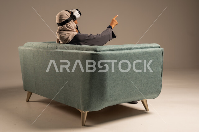 قضاء أوقات ممتعة ومسلية بالألعاب الافتراضية، مشاهدة أفلام الواقع الافتراضي، وسائل ترفيهية في وقت الفراغ,  بورتريه لإمرأة عربية خليجية سعودية تجلس على كرسي مريح وترتدي نظارة ثلاثية الأبعاد، خلفية رمادية