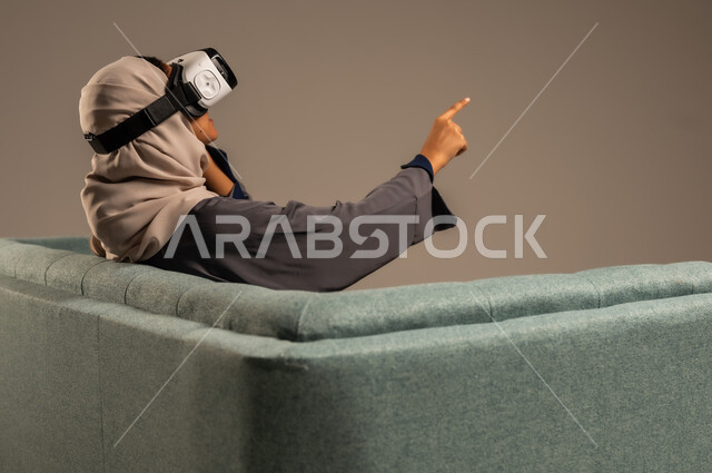 قضاء أوقات ممتعة ومسلية بالألعاب الافتراضية، مشاهدة أفلام الواقع الافتراضي، وسائل ترفيهية في وقت الفراغ,  بورتريه لإمرأة عربية خليجية سعودية تجلس على كرسي مريح وترتدي نظارة ثلاثية الأبعاد، خلفية رمادية