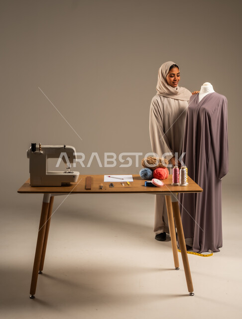 ممارسة مهنة الخياطة , بورتريه لمصممة أزياء عربية خليجية ترتدي العباءة الملونة والحجاب تقف في ورشة عمل تصميم أزياء, حرف و مهن يدوية في المملكة، أعمال تقليدية شعبية, اتقان صنع الملابس السعودية، الطول الكامل للجسم، خلفية رمادية