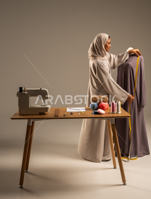ممارسة مهنة الخياطة , بورتريه لمصممة أزياء عربية خليجية ترتدي العباءة الملونة والحجاب تقف في ورشة عمل تصميم أزياء, حرف و مهن يدوية في المملكة، أعمال تقليدية شعبية, اتقان صنع الملابس السعودية، الطول الكامل للجسم، خلفية رمادية