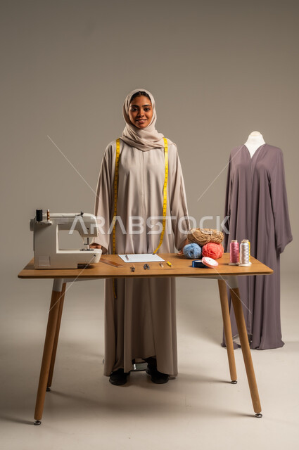 ممارسة مهنة الخياطة , بورتريه لمصممة أزياء عربية خليجية ترتدي العباءة الملونة والحجاب تقف في ورشة عمل تصميم أزياء, حرف و مهن يدوية في المملكة، أعمال تقليدية شعبية, اتقان صنع الملابس السعودية، الطول الكامل للجسم، خلفية رمادية