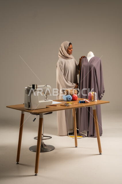 ممارسة مهنة الخياطة , بورتريه لمصممة أزياء عربية خليجية ترتدي العباءة الملونة والحجاب تقف في ورشة عمل تصميم أزياء, حرف و مهن يدوية في المملكة، أعمال تقليدية شعبية, اتقان صنع الملابس السعودية، الطول الكامل للجسم، خلفية رمادية