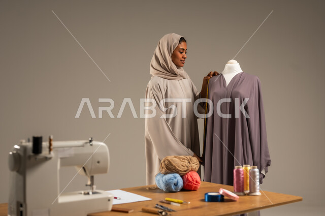 Practicing the profession of sewing, portrait of an Arab Gulf fashion designer wearing a colorful abaya and hijab standing in a fashion design workshop, crafts and handicrafts in the Kingdom, traditional folk works, mastery of making Saudi clothes, full body length, gray background