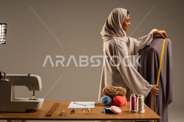 أعمال تقليدية شعبية, ممارسة مهنة الخياطة , بورتريه لمصممة أزياء عربية خليجية ترتدي العباءة الملونة والحجاب تقف في ورشة عمل تصميم أزياء, حرف و مهن يدوية في المملكة، اتقان صنع الملابس السعودية، خلفية رمادية