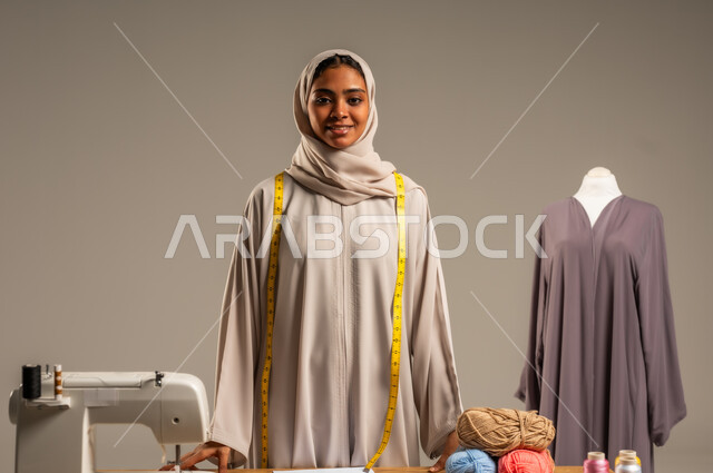 أعمال تقليدية شعبية, ممارسة مهنة الخياطة , بورتريه لمصممة أزياء عربية خليجية ترتدي العباءة الملونة والحجاب تقف في ورشة عمل تصميم أزياء, حرف و مهن يدوية في المملكة، اتقان صنع الملابس السعودية، خلفية رمادية