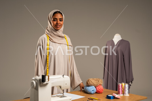 أعمال تقليدية شعبية, ممارسة مهنة الخياطة , بورتريه لمصممة أزياء عربية خليجية ترتدي العباءة الملونة والحجاب تقف في ورشة عمل تصميم أزياء, حرف و مهن يدوية في المملكة، اتقان صنع الملابس السعودية، خلفية رمادية