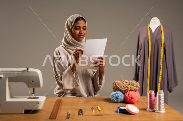 Crafts and handicrafts in the Kingdom, traditional folk works, practicing the profession of sewing, portrait of a Gulf Arab fashion designer wearing a colorful abaya and hijab drawing on a white sheet of paper in a fashion design workshop, mastering the making of Saudi clothes, gray background