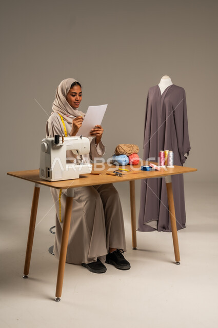 Crafts and handicrafts in the Kingdom, traditional folk works, practicing the profession of sewing, portrait of a Gulf Arab fashion designer wearing a colorful abaya and hijab drawing on a white sheet of paper in a fashion design workshop, mastering the making of Saudi clothes, gray background