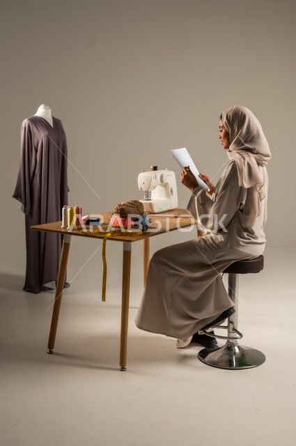 Crafts and handicrafts in the Kingdom, traditional folk works, practicing the profession of sewing, portrait of a Gulf Arab fashion designer wearing a colorful abaya and hijab drawing on a white sheet of paper in a fashion design workshop, mastering the making of Saudi clothes, gray background