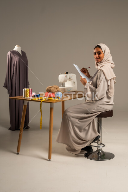 Crafts and handicrafts in the Kingdom, traditional folk works, practicing the profession of sewing, portrait of a Gulf Arab fashion designer wearing a colorful abaya and hijab drawing on a white sheet of paper in a fashion design workshop, mastering the making of Saudi clothes, gray background