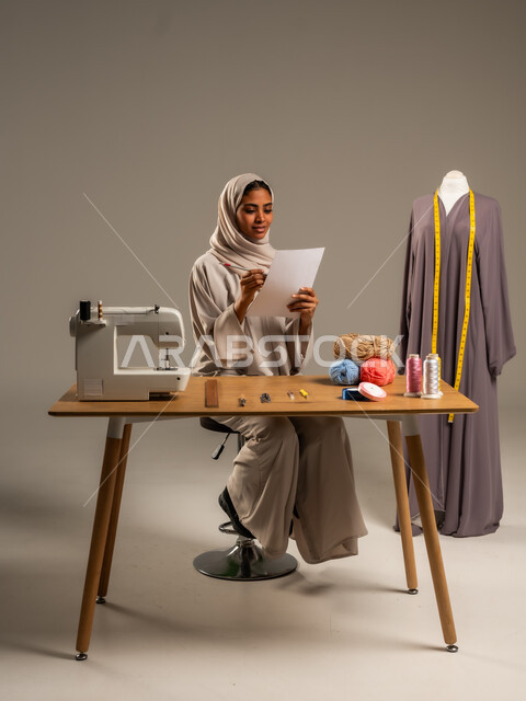 Professions and handicrafts in the Kingdom, traditional folk works, practicing the profession of sewing, portrait of an Arab Gulf fashion designer wearing a colorful abaya and hijab drawing on a white sheet of paper in a fashion design workshop, mastering the making of Saudi clothes, gray background