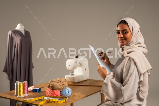  مهن وحرف يدوية في المملكة، أعمال تقليدية شعبية, ممارسة مهنة الخياطة , بورتريه لمصممة أزياء عربية خليجية ترتدي العباءة الملونة والحجاب تقوم بالرسم على ورقة بيضاء في ورشة عمل تصميم أزياء, اتقان صنع الملابس السعودية، خلفية رمادية
