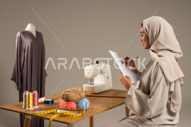  مهن وحرف يدوية في المملكة، أعمال تقليدية شعبية, ممارسة مهنة الخياطة , بورتريه لمصممة أزياء عربية خليجية ترتدي العباءة الملونة والحجاب تقوم بالرسم على ورقة بيضاء في ورشة عمل تصميم أزياء, اتقان صنع الملابس السعودية، خلفية رمادية