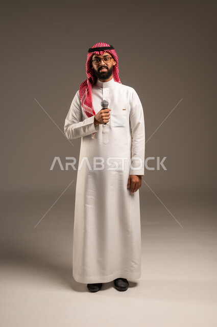 تقديم العروضات المسرحية، الاستمتاع بأداء المواهب الصوتية، بورتريه لشاب عربي خليجي سعودي يرتدي الثوب التقليدي والشماغ يمسك بيده المايكروفون، إيماءات الاندماج والتفاعل مع الموسيقا، تسجيل مقطع صوتي غنائي، خلفيه رمادية