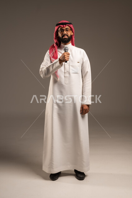 تقديم العروضات المسرحية، الاستمتاع بأداء المواهب الصوتية، بورتريه لشاب عربي خليجي سعودي يرتدي الثوب التقليدي والشماغ يمسك بيده المايكروفون، إيماءات الاندماج والتفاعل مع الموسيقا، تسجيل مقطع صوتي غنائي، خلفيه رمادية