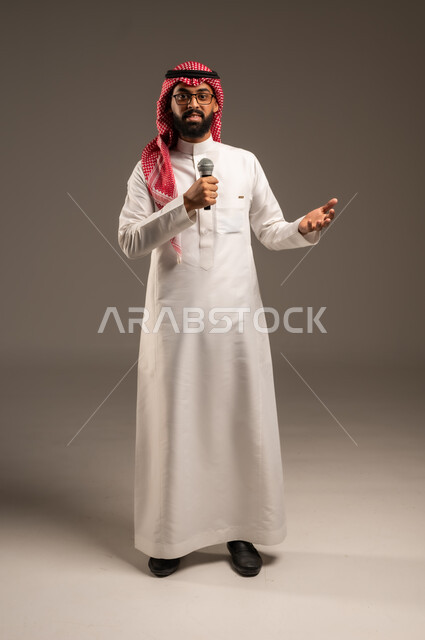 تقديم العروضات المسرحية، الاستمتاع بأداء المواهب الصوتية، بورتريه لشاب عربي خليجي سعودي يرتدي الثوب التقليدي والشماغ يمسك بيده المايكروفون، إيماءات الاندماج والتفاعل مع الموسيقا، تسجيل مقطع صوتي غنائي، خلفيه رمادية