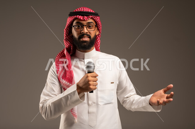 تقديم العروضات المسرحية، الاستمتاع بأداء المواهب الصوتية، بورتريه لشاب عربي خليجي سعودي يرتدي الثوب التقليدي والشماغ يمسك بيده المايكروفون، إيماءات الاندماج والتفاعل مع الموسيقا، تسجيل مقطع صوتي غنائي، خلفيه رمادية