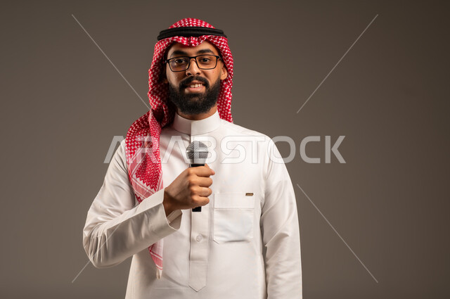 تقديم العروضات المسرحية، الاستمتاع بأداء المواهب الصوتية، بورتريه لشاب عربي خليجي سعودي يرتدي الثوب التقليدي والشماغ يمسك بيده المايكروفون، إيماءات الاندماج والتفاعل مع الموسيقا، تسجيل مقطع صوتي غنائي، خلفيه رمادية