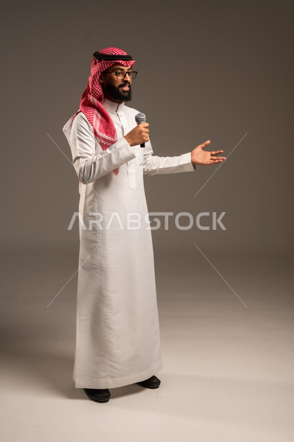 تقديم العروضات المسرحية، الاستمتاع بأداء المواهب الصوتية، بورتريه لشاب عربي خليجي سعودي يرتدي الثوب التقليدي والشماغ يمسك بيده المايكروفون، إيماءات الاندماج والتفاعل مع الموسيقا، تسجيل مقطع صوتي غنائي، خلفيه رمادية
